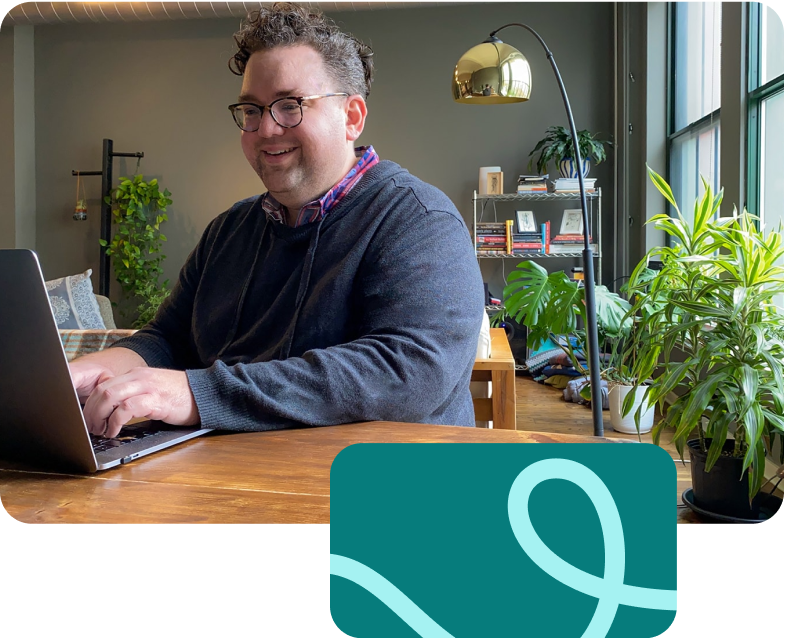 A male-identifying engineer smiles in his home office while working on his laptop.