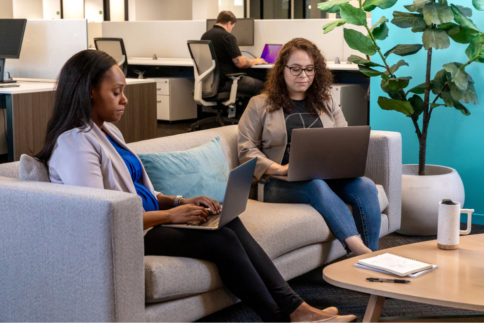 Two female-identifying Sprout employees sit comfortably on a couch, concentrating as they work on their laptops.
