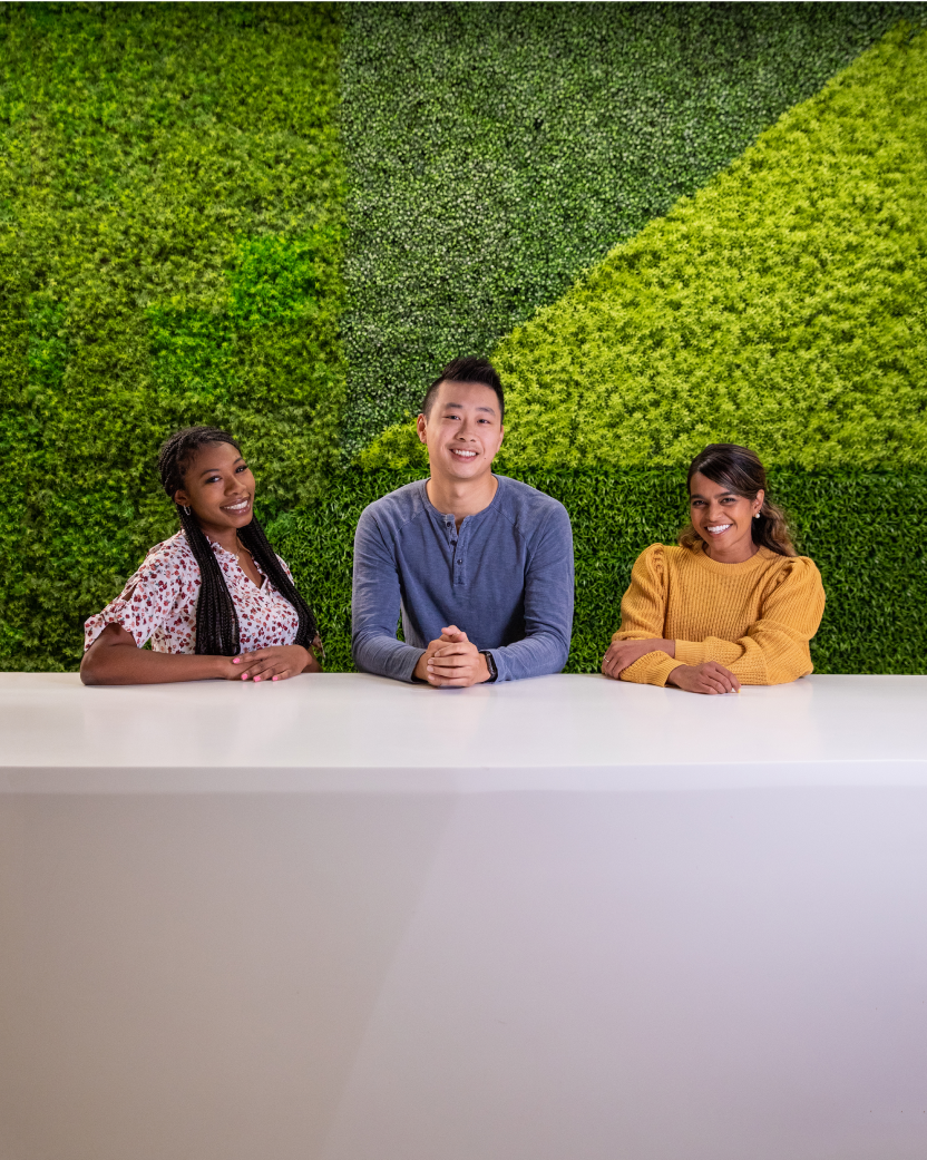 Racially and gender diverse Sprout employees smiling while posing in front of a bright green grass wall the office.